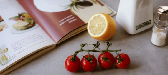 Flat lay of cooking ingredients including tomatoes and a lemon with a cookbook.